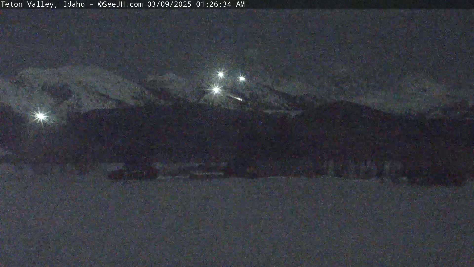 A nighttime, snowy landscape shows a dark mountain range with a few bright lights visible in the distance.