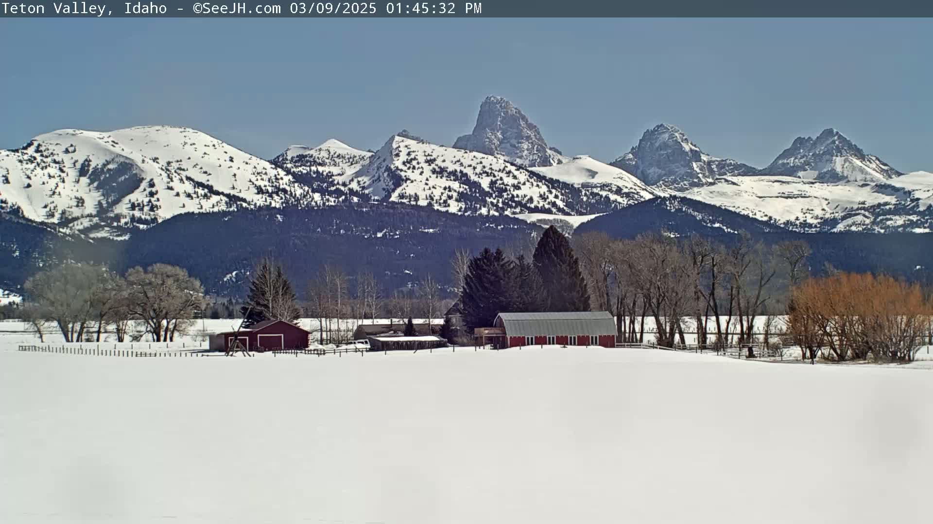A snow-covered landscape features a red barn and other farm buildings nestled among leafless trees, with snow-capped mountains in the background under a clear sky.