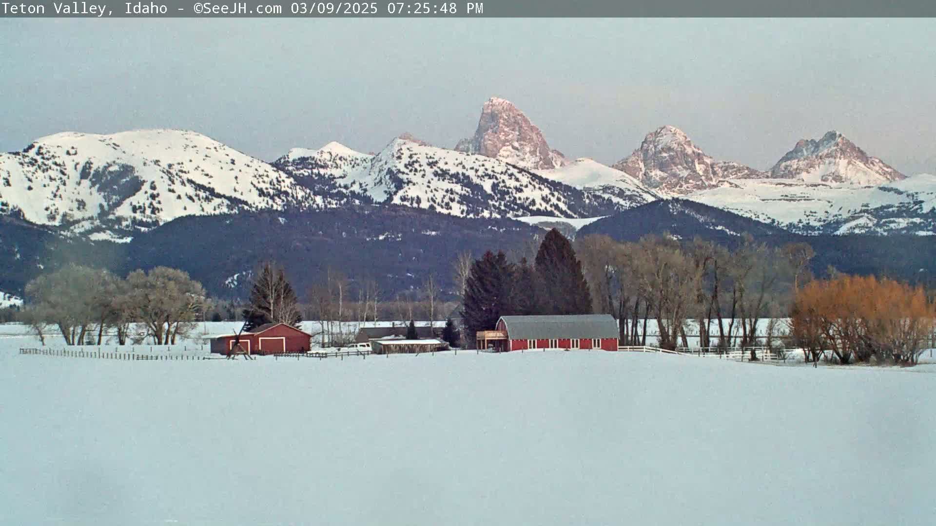 A red barn sits on a snow-covered field in front of snow-capped mountains under a clear sky.