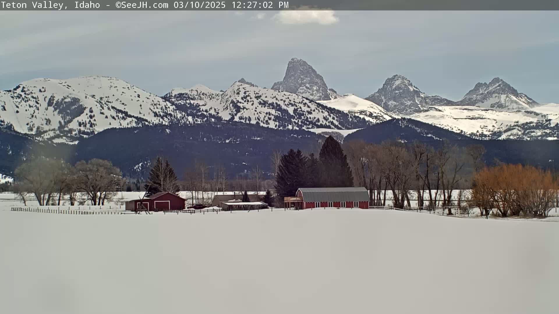 A snow-covered landscape features a red barn and other buildings, situated in front of a range of snow-capped mountains under a mostly cloudy sky.