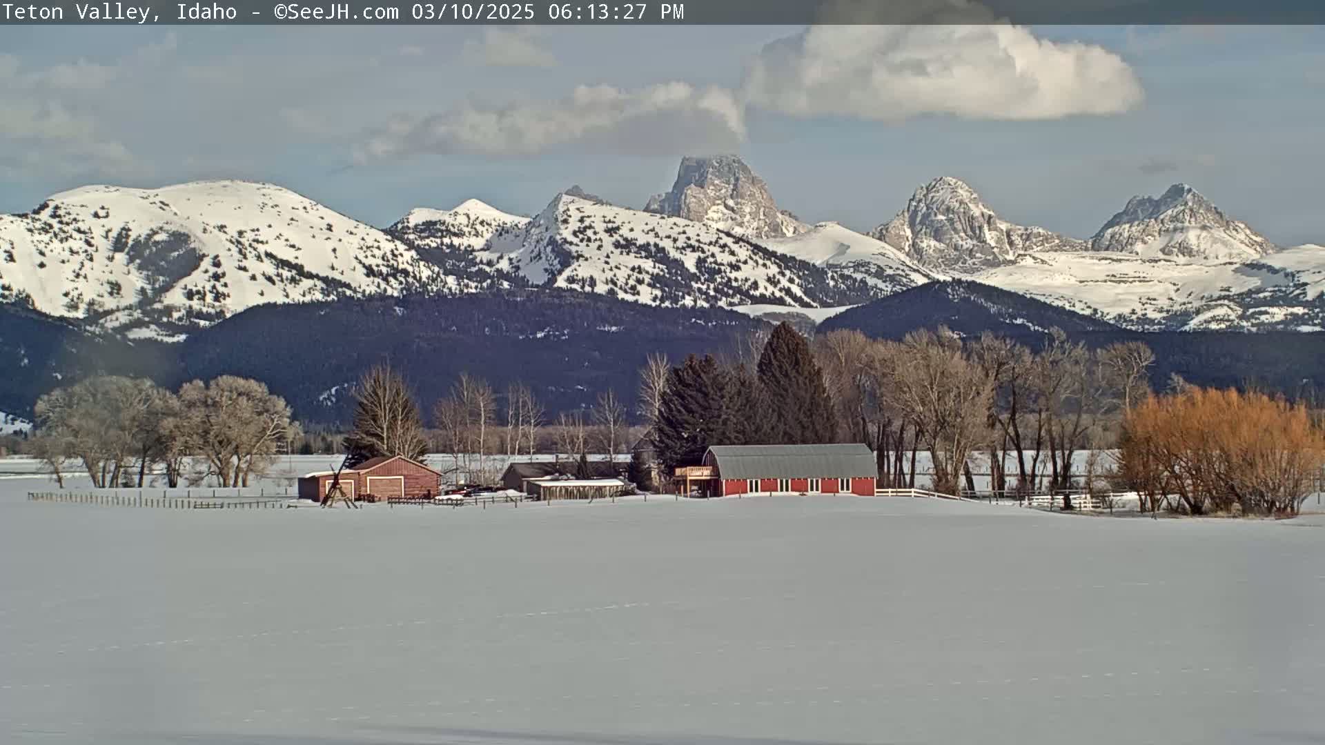 A snow-covered field with a red barn and other buildings sits in front of a range of snow-capped mountains under a partly cloudy sky.