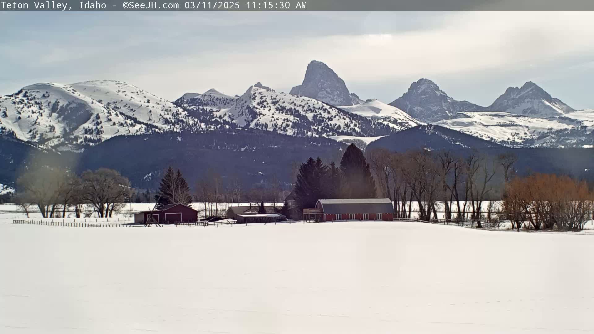 A snow-covered valley with a red barn and other buildings, set against a backdrop of snow-capped mountains under a partly cloudy sky.