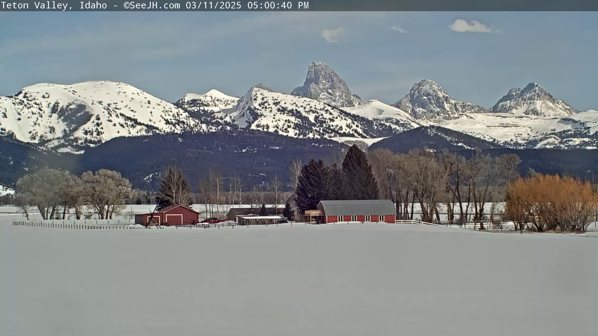 A snow-covered landscape features a red barn and other farm buildings nestled amongst leafless trees in the foreground, with snow-capped mountains rising in the background under a mostly clear sky.