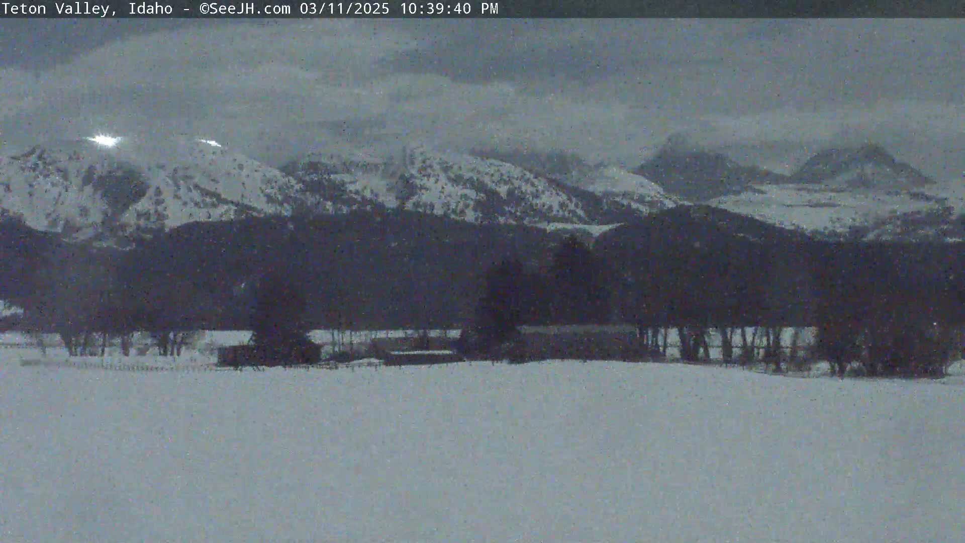 A snow-covered landscape features a line of trees and buildings in the foreground, with snow-capped mountains in the background under an overcast sky.
