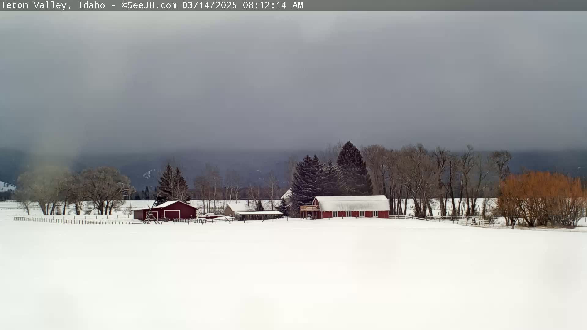 A snow-covered landscape features a red barn and other outbuildings, surrounded by leafless trees under an overcast sky.