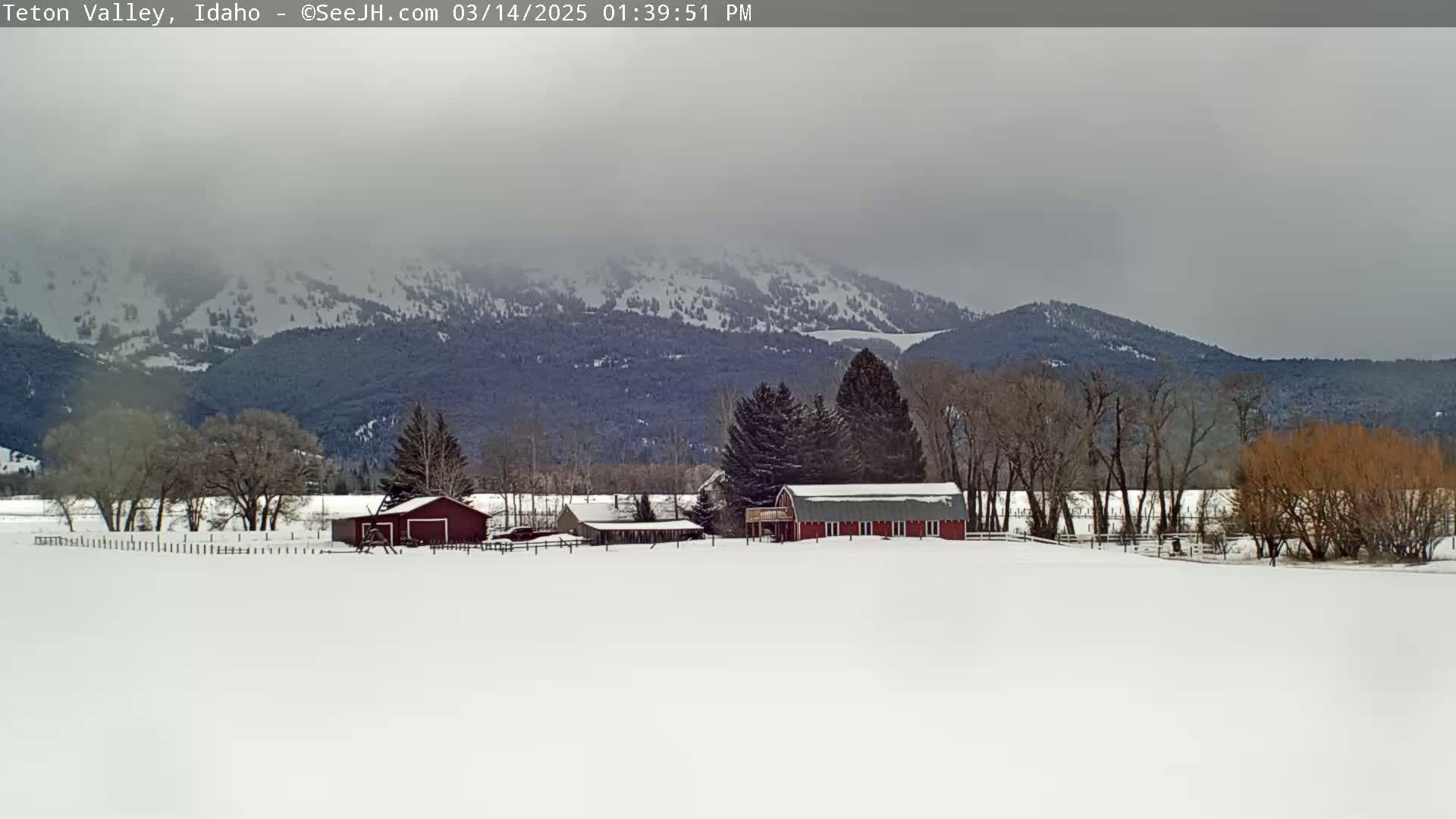 A snow-covered landscape features a red barn and other farm buildings nestled amongst leafless trees, with a snow-capped mountain range in the background under an overcast sky.