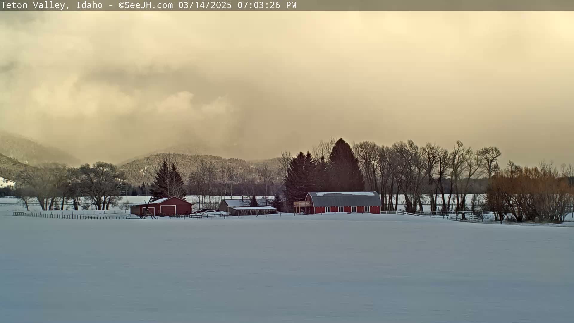 A snow-covered landscape features a red barn and other farm buildings, surrounded by leafless trees and a distant mountain range under a hazy, light-colored sky.