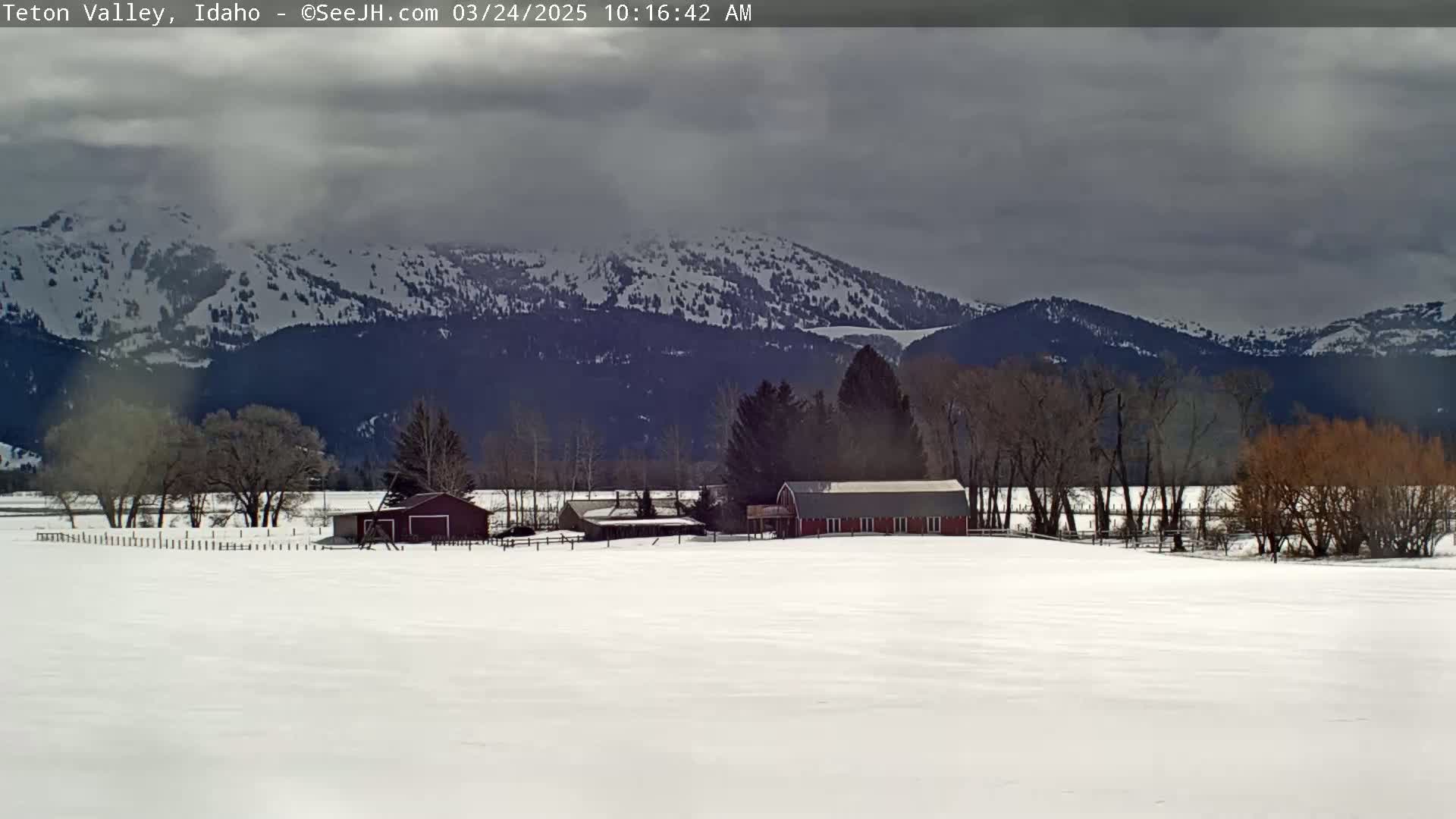 A snow-covered field with a red barn and outbuildings sits in front of a mountain range under a cloudy sky.