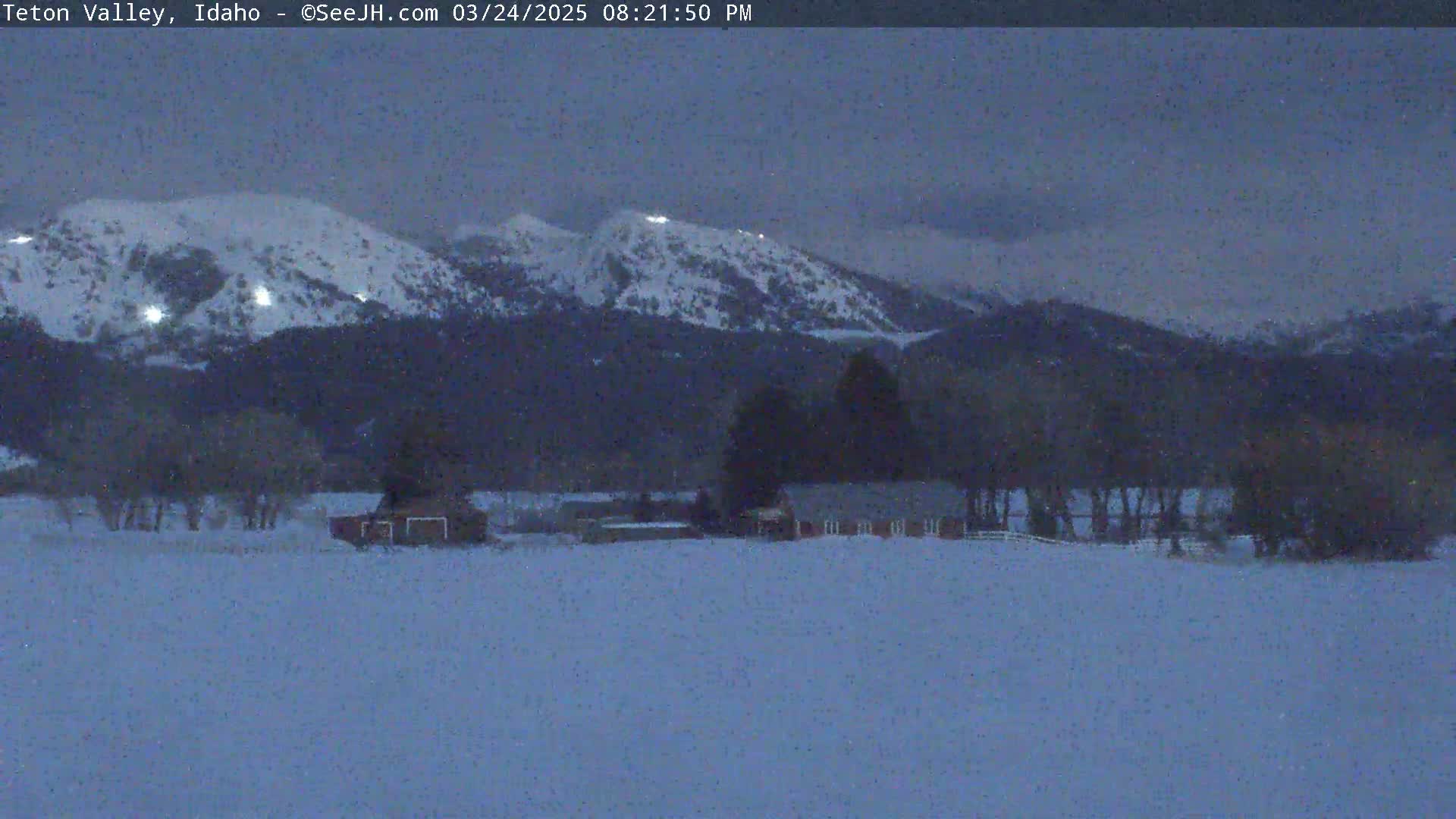 Snow-covered mountains are visible in the distance under a twilight sky, with a snow-covered field and several buildings in the foreground.
