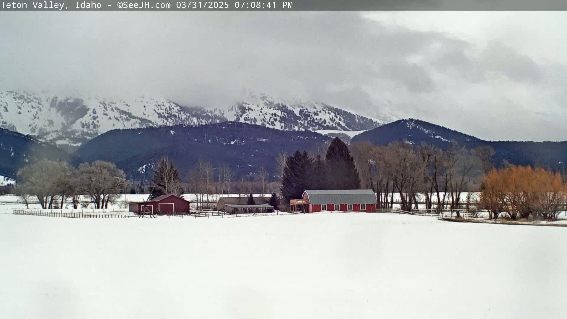 A snow-covered field with a red barn and other farm buildings sits in front of a mountain range under an overcast sky.