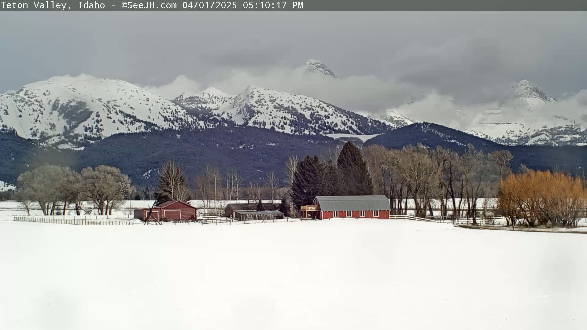 A snow-covered valley with a red barn and other farm buildings, sits in front of a range of snow-capped mountains under a cloudy sky.