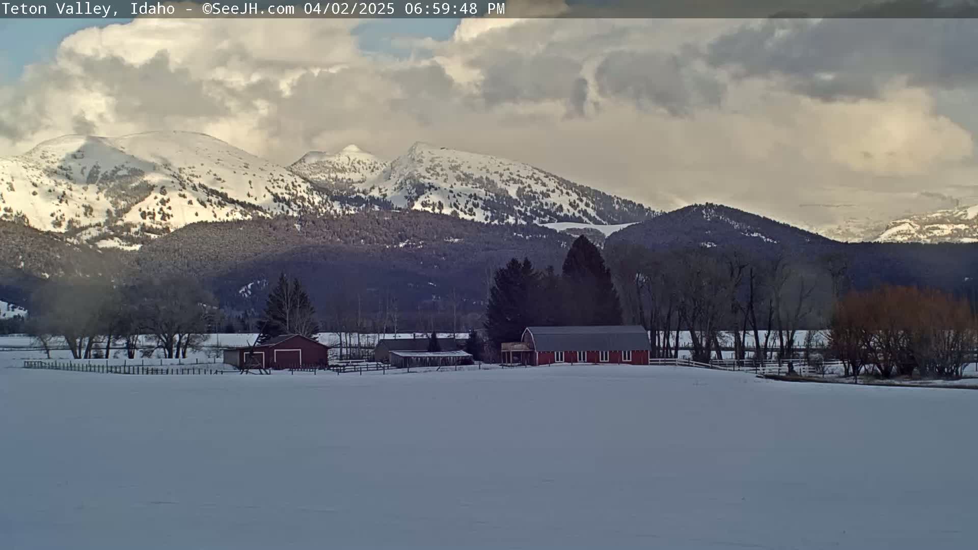 A snow-covered valley with a red barn and outbuildings, surrounded by leafless trees, sits before a range of snow-capped mountains under a partly cloudy sky.