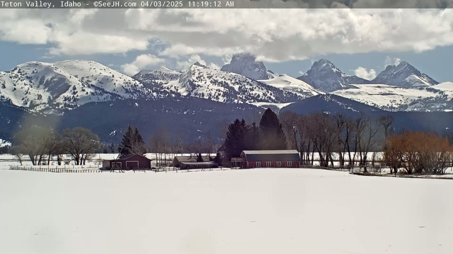 A snow-covered valley with a red barn and other farm buildings is nestled beneath a partly cloudy sky and a range of snow-capped mountains.