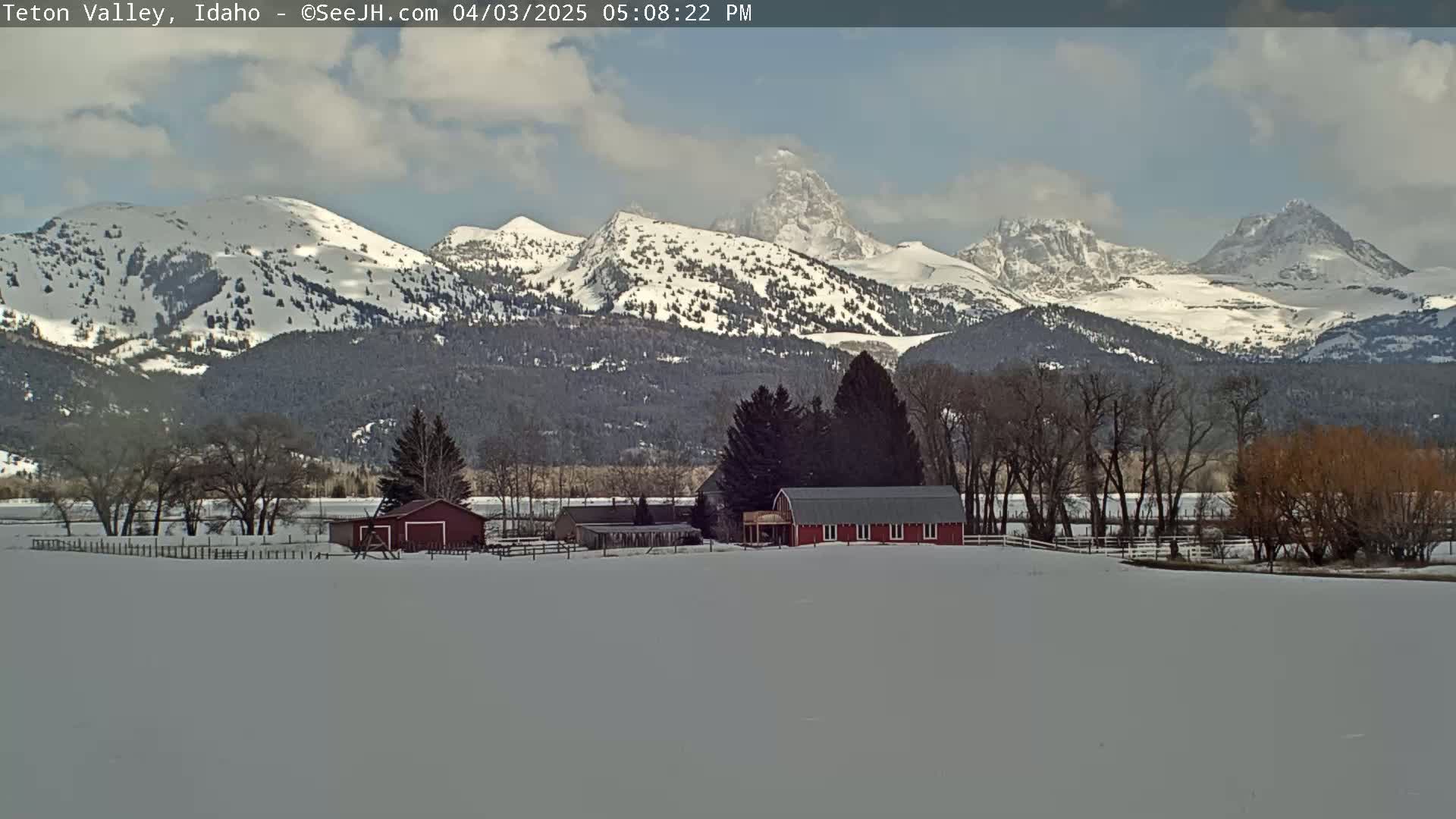 A snow-covered landscape features a red barn and other farm structures in the foreground, with snow-capped mountains rising in the background under a partly cloudy sky.