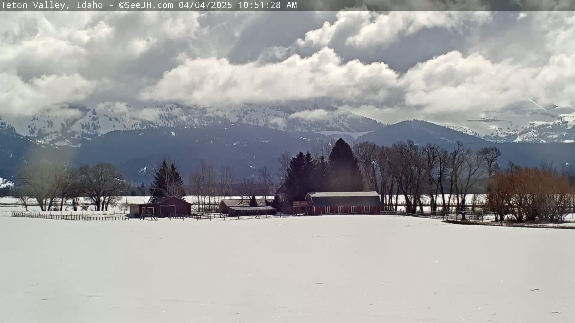 A snow-covered field with a red barn and other buildings sits in front of a mountain range under a partly cloudy sky.