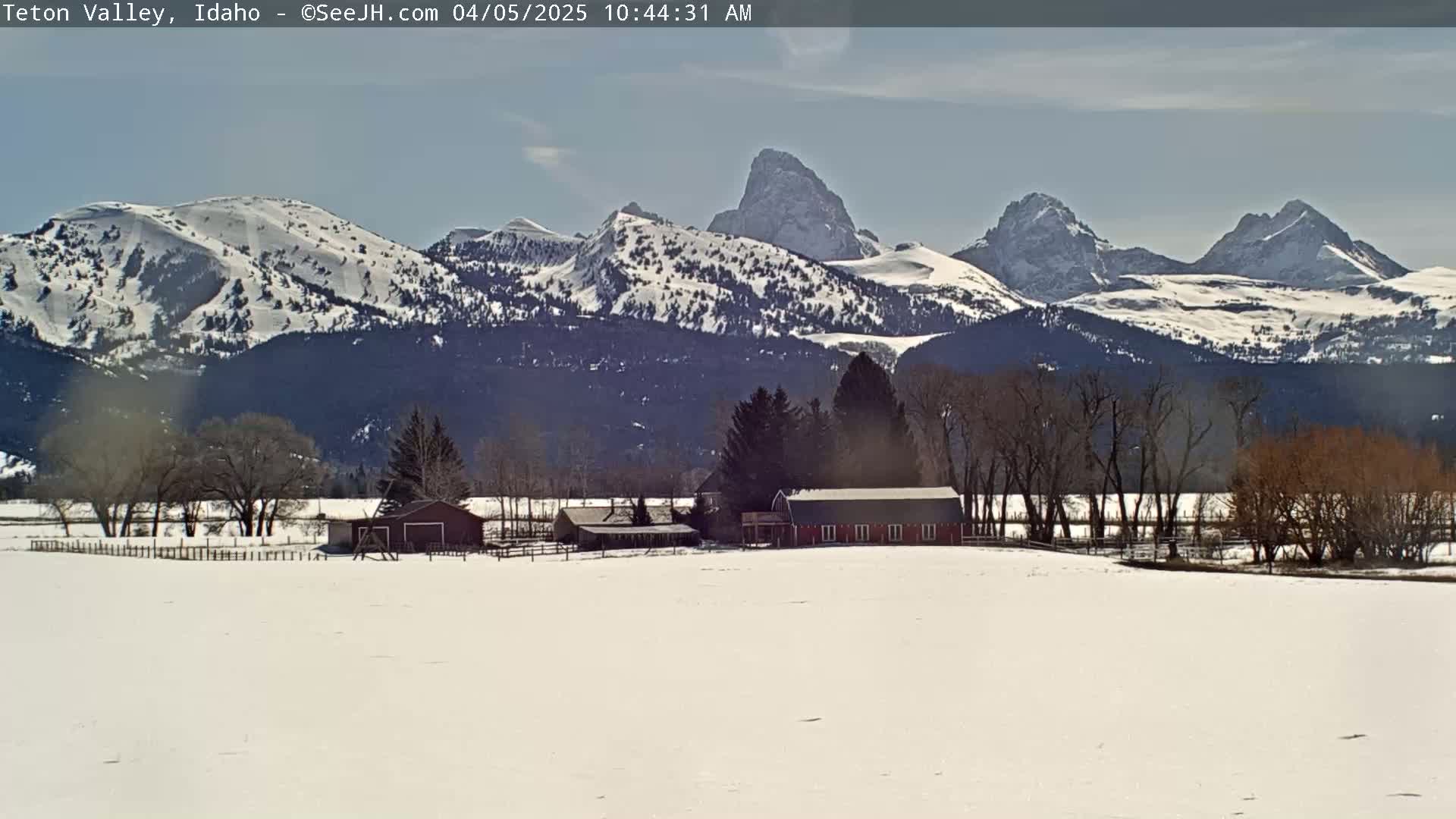 A snow-covered field with a red barn and other buildings sits in the foreground of a bright, sunny day with a range of snow-capped mountains in the background.