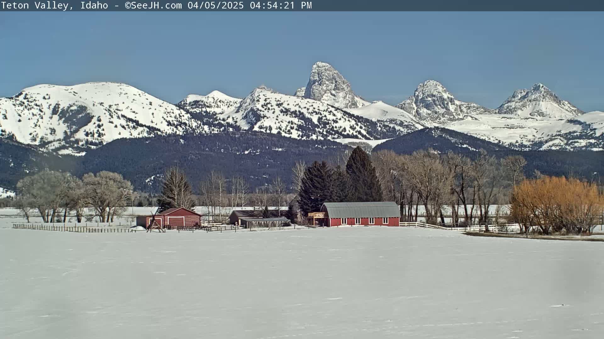 A snow-covered valley with a red barn and outbuildings sits in the foreground, with snow-capped mountains rising in the background under a clear blue sky.