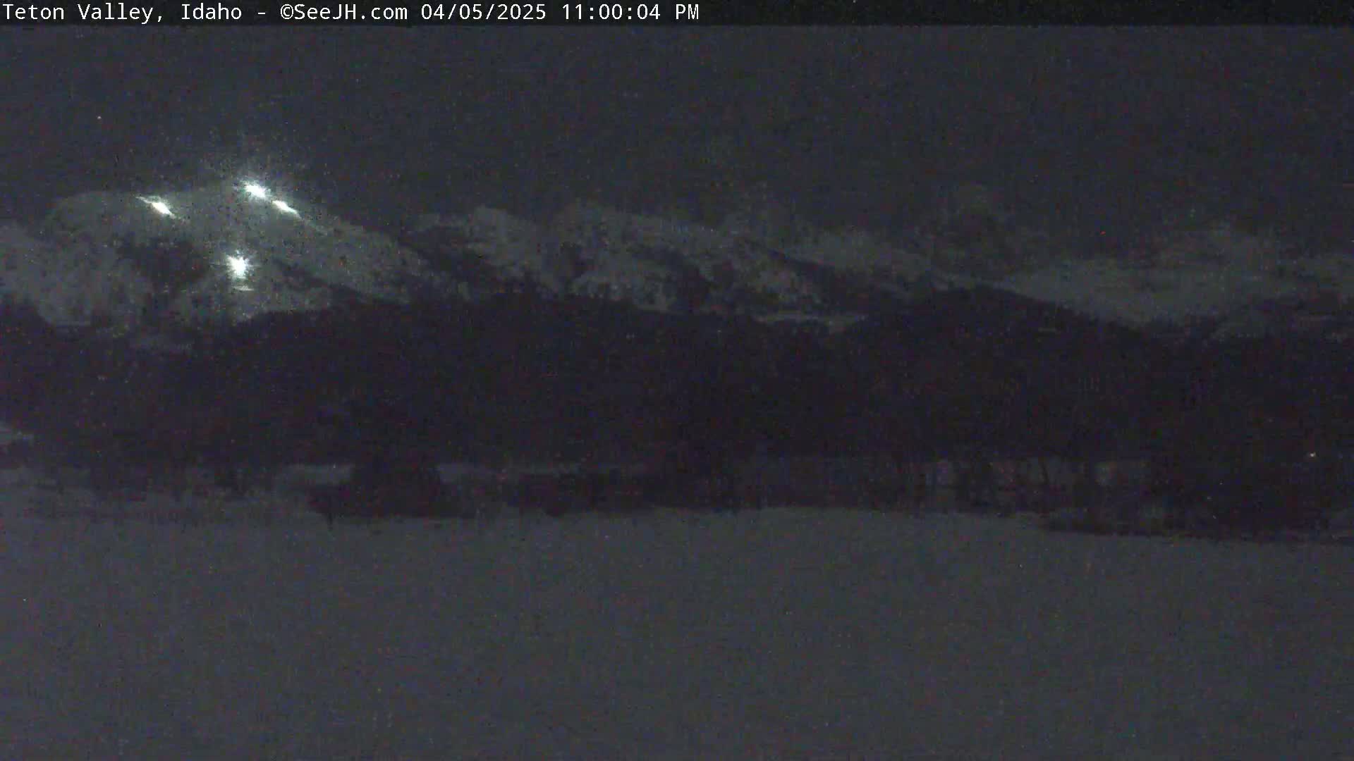 A nighttime, snow-covered landscape shows a mountain range silhouetted against a dark sky, illuminated by several bright lights in the distance.