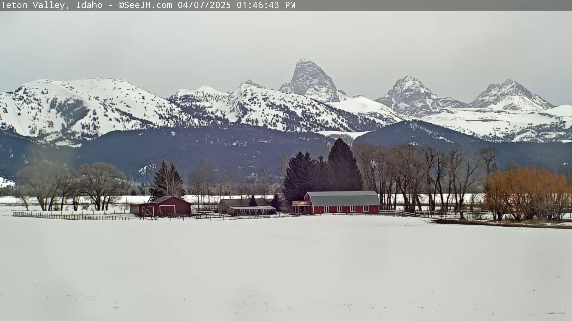 A snow-covered valley features a red barn and outbuildings, with snow-capped mountains in the background under an overcast sky.