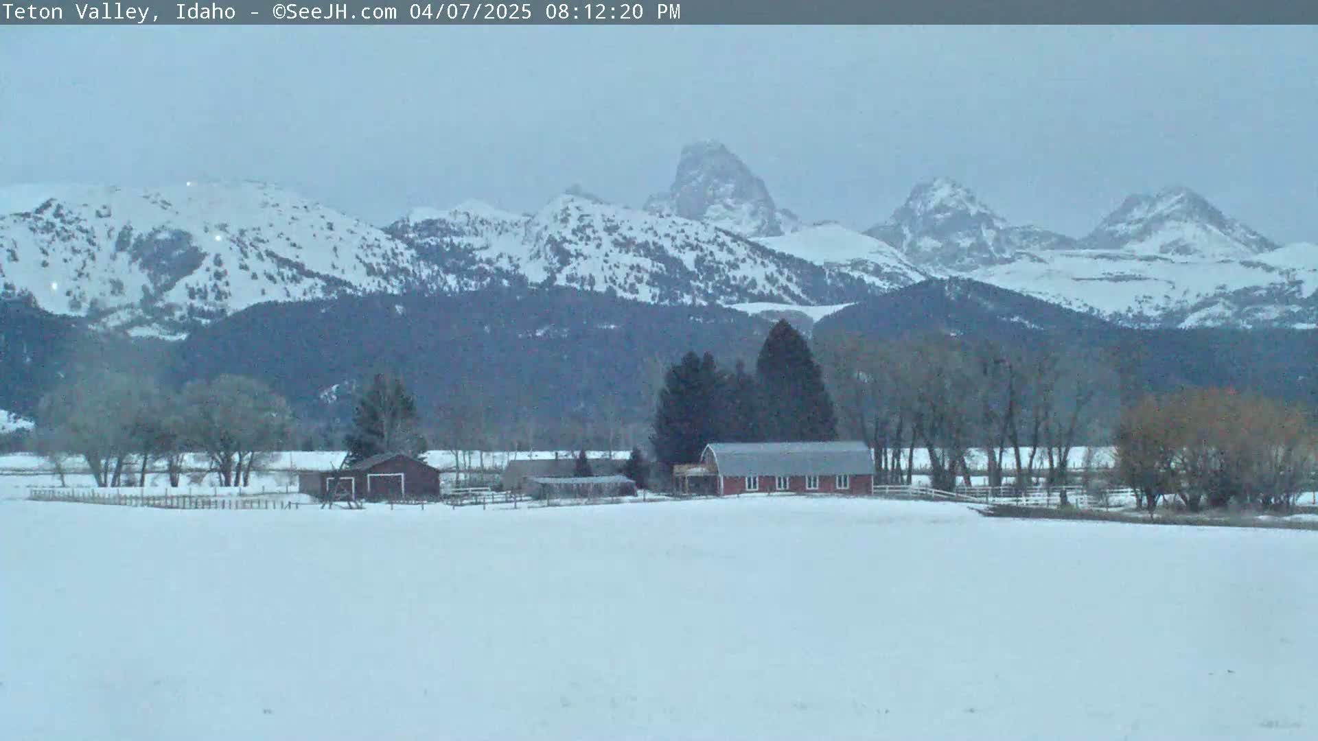 A snow-covered valley with a red barn and outbuildings sits in the foreground of a mountain range under an overcast sky.