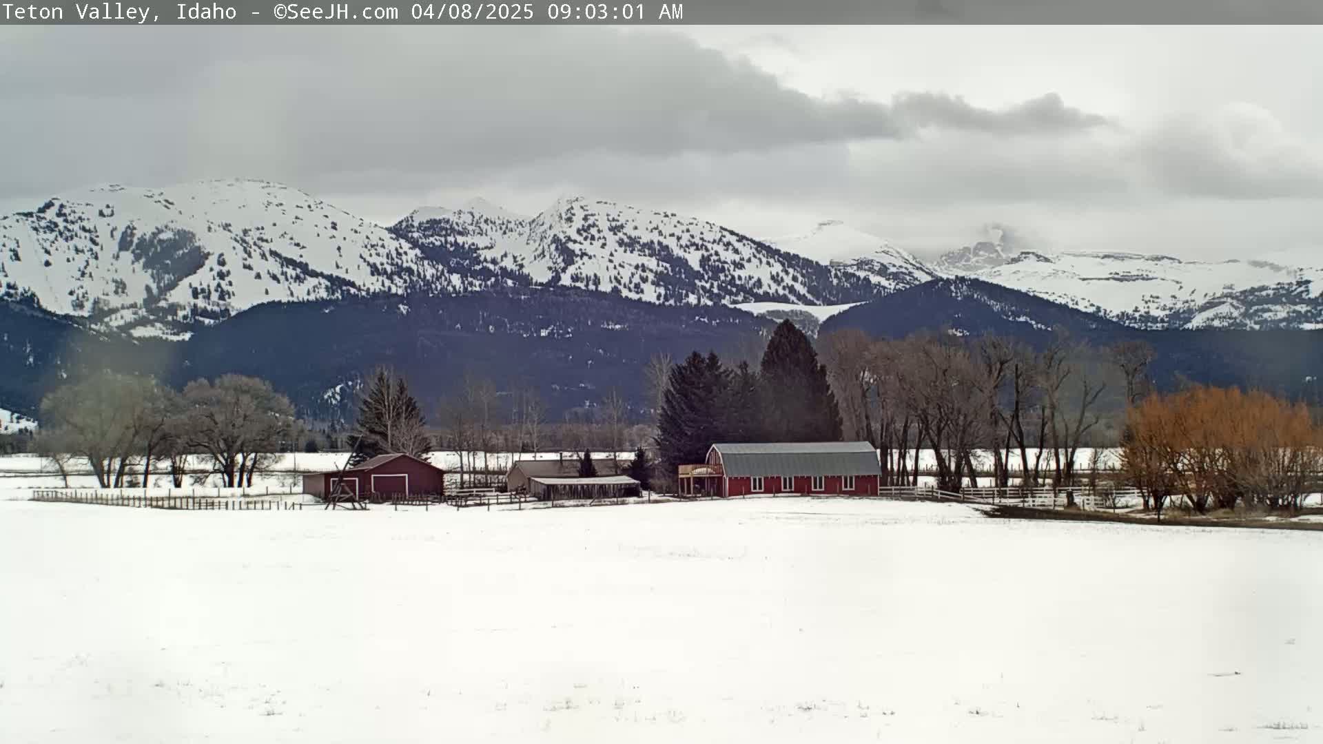 A snow-covered landscape features a red barn and other farm buildings, with snow-capped mountains in the background under an overcast sky.