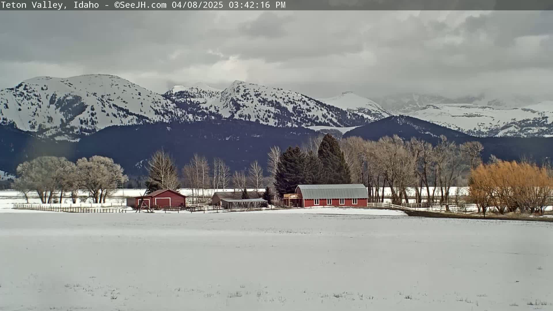 A snow-covered landscape features a red barn and other farm buildings nestled among leafless trees, with a backdrop of snow-capped mountains under a cloudy sky.