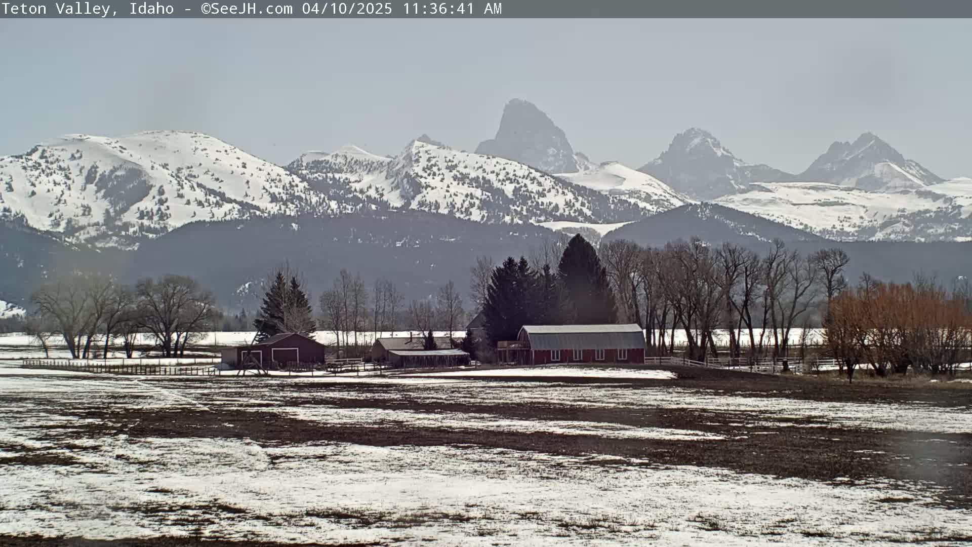 A farm with a red barn sits in a snow-dusted field, overlooking snow-capped mountains under a mostly clear sky.