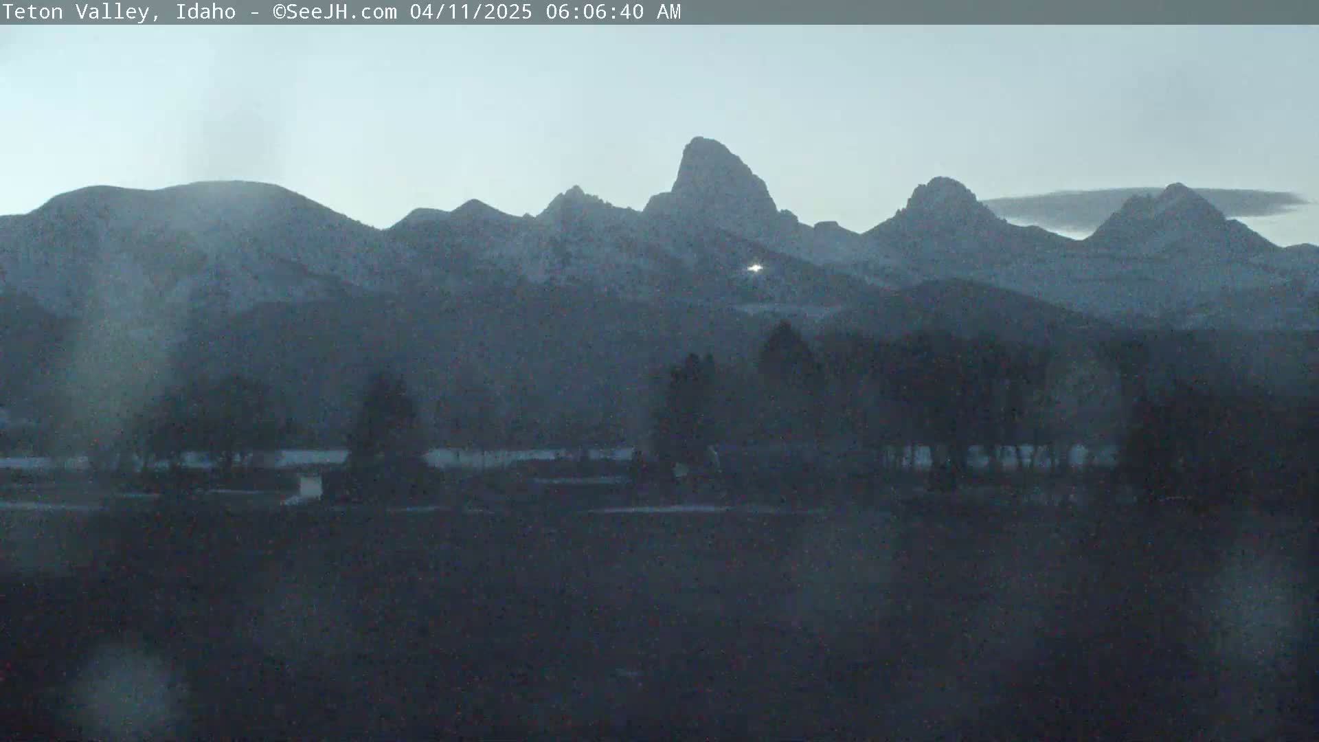 Snow-dusted mountains rise in the distance under a twilight sky, with a dark foreground of trees and flat land.