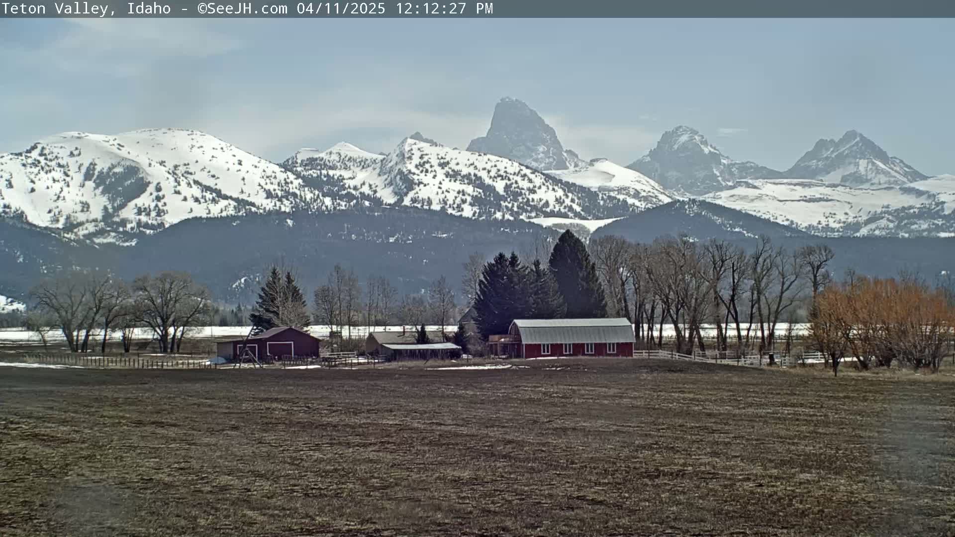 A red barn sits on a farm in front of snow-capped mountains under a mostly clear sky.