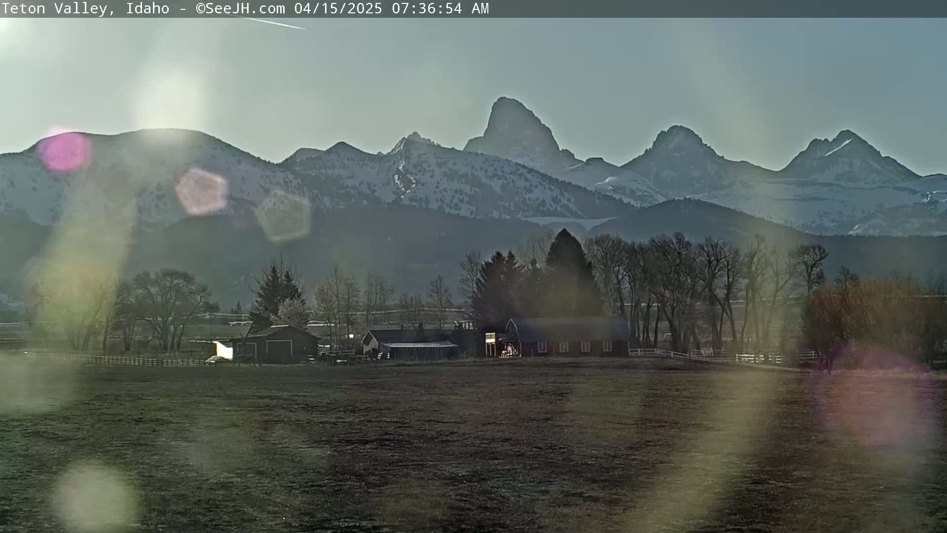 A farm with a red barn sits in a field before snow-capped mountains under a mostly clear, bright sky.
