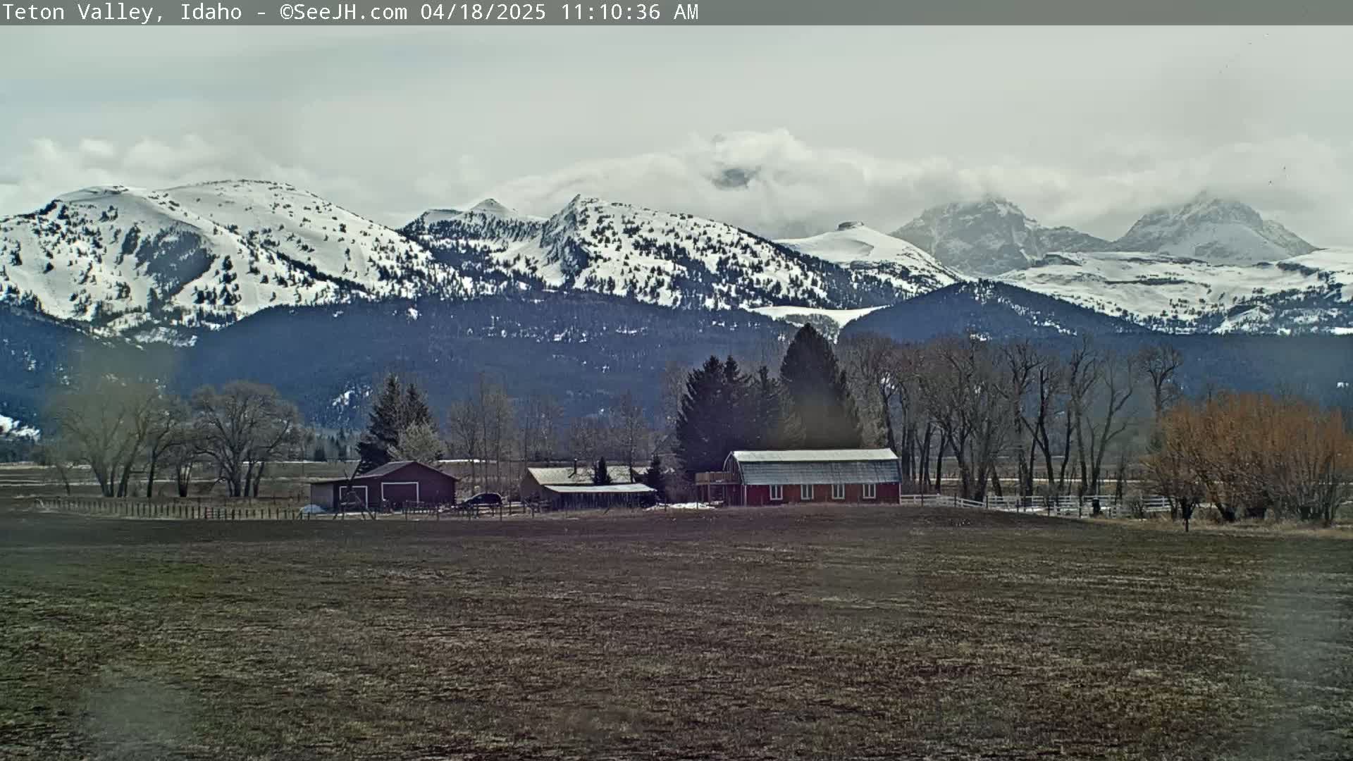 A farm with a red barn sits in a field before a range of snow-capped mountains under a cloudy sky.