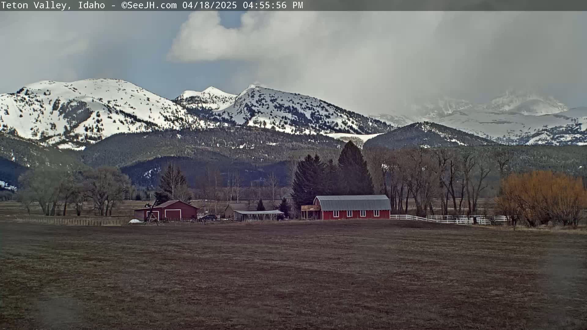 A red barn sits on a flat, brown field in front of snow-capped mountains under a cloudy sky.