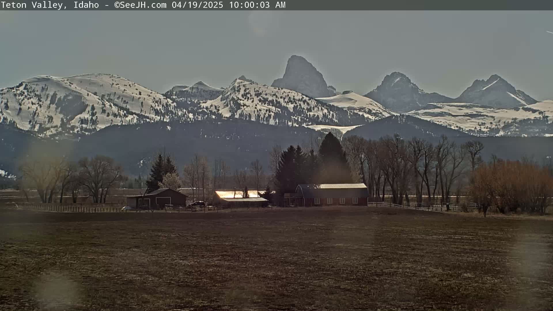 A farm with several buildings sits in a valley before snow-capped mountains under a mostly clear sky.