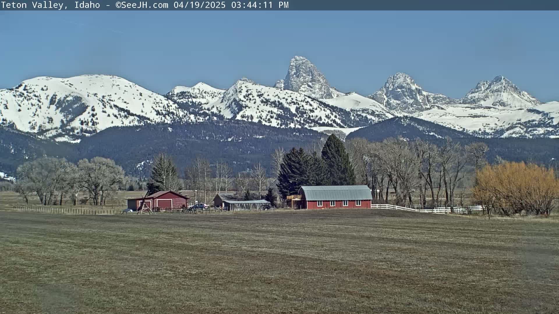 A snow-capped mountain range rises behind a farm with a red barn and outbuildings on a clear, sunny day.