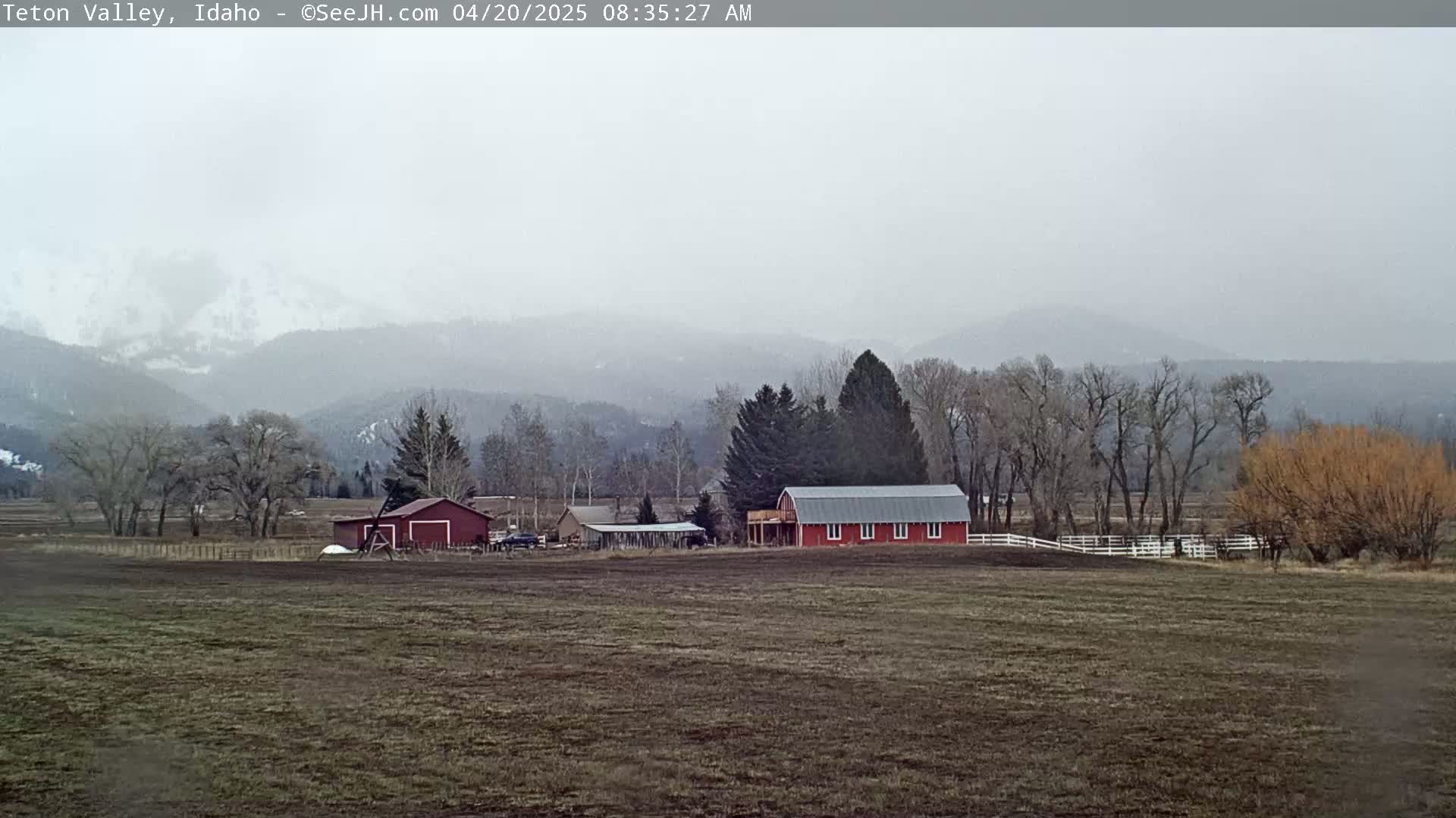 A hazy, overcast day reveals a farm with red barns and outbuildings in a field, situated against a backdrop of distant, snow-dusted mountains.