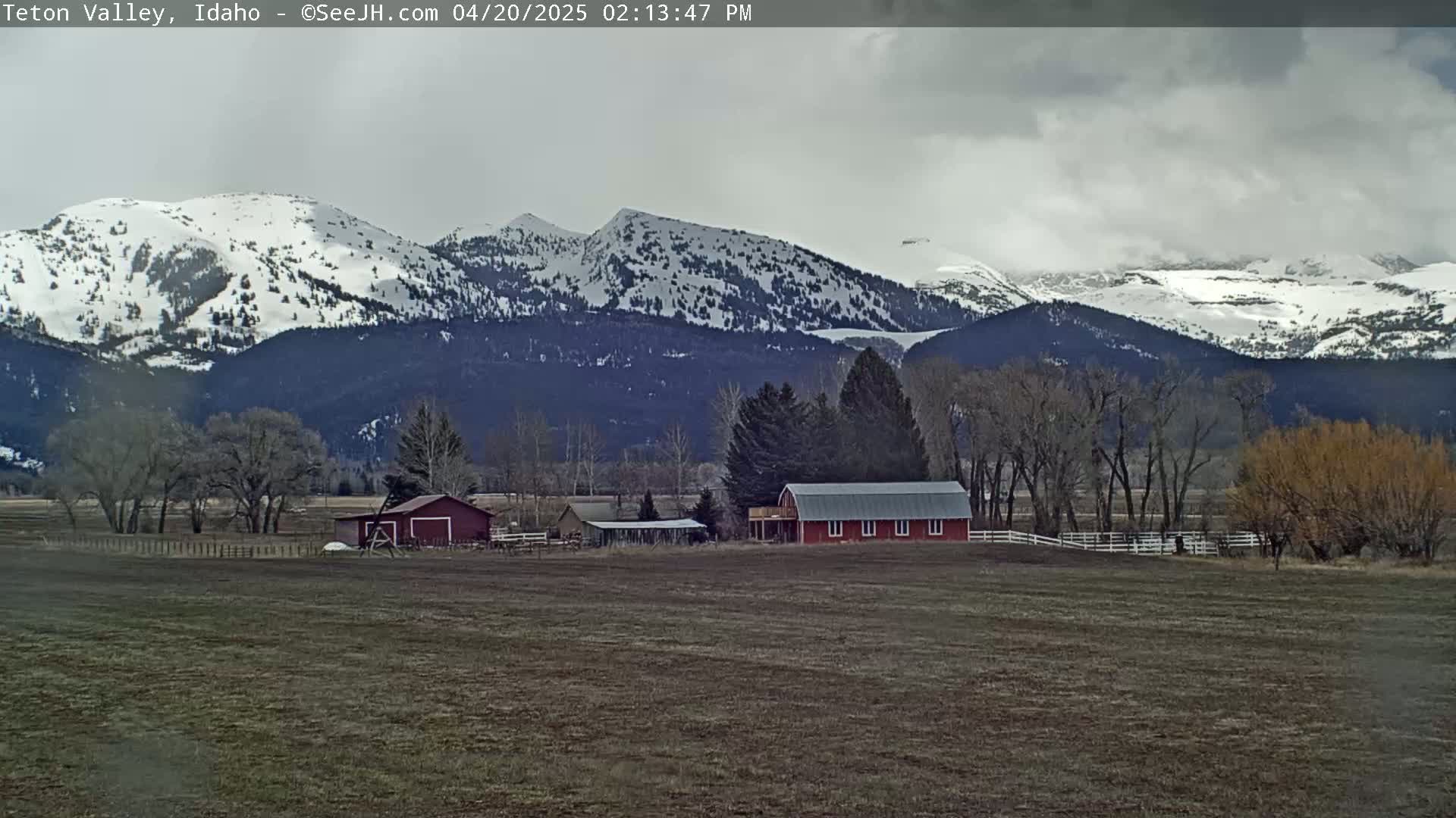 A red barn sits on a farm in a valley, with snow-capped mountains in the background under a cloudy sky.