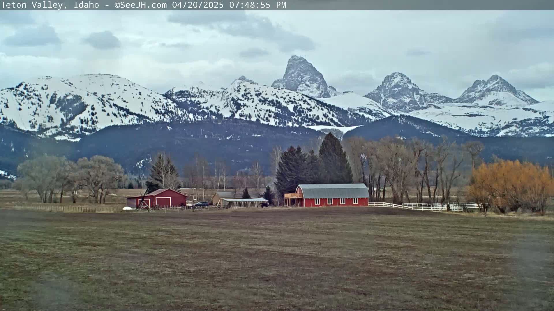 A flat, brown field stretches out before a red barn and a cluster of trees, all set against a backdrop of snow-capped mountains under a cloudy sky.