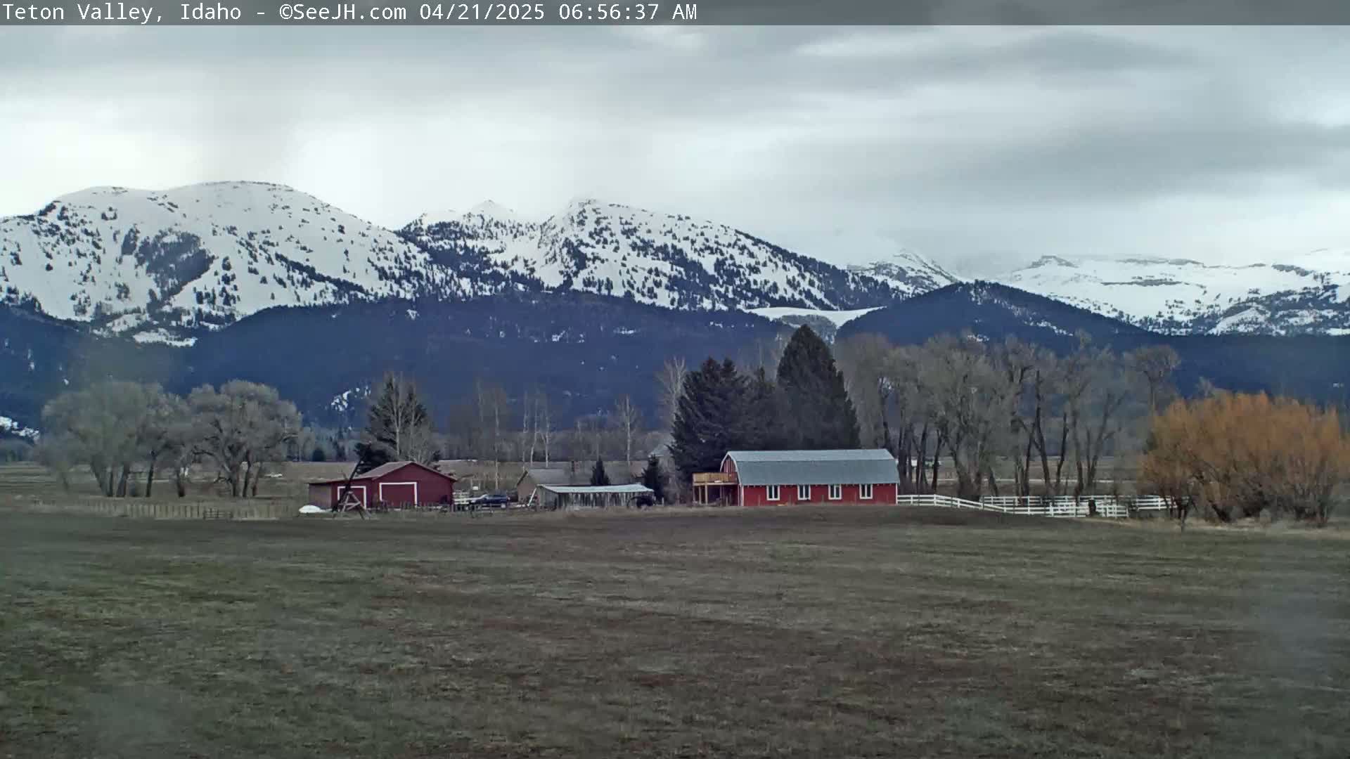 A snow-capped mountain range is visible in the background of a flat, field with a red barn and other buildings, under an overcast sky.