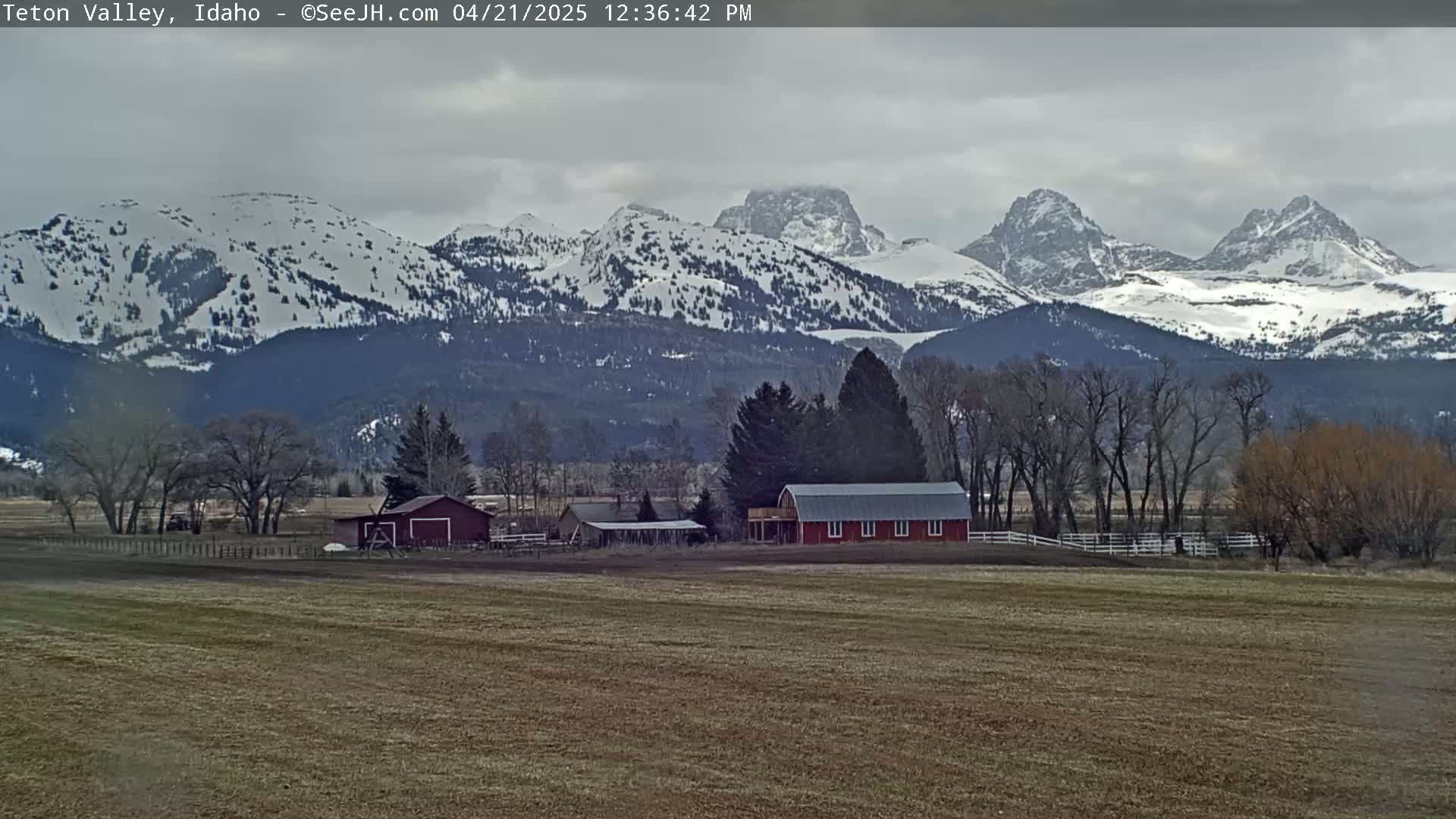 A snow-capped mountain range rises behind a flat field containing a red barn and other farm buildings under an overcast sky.