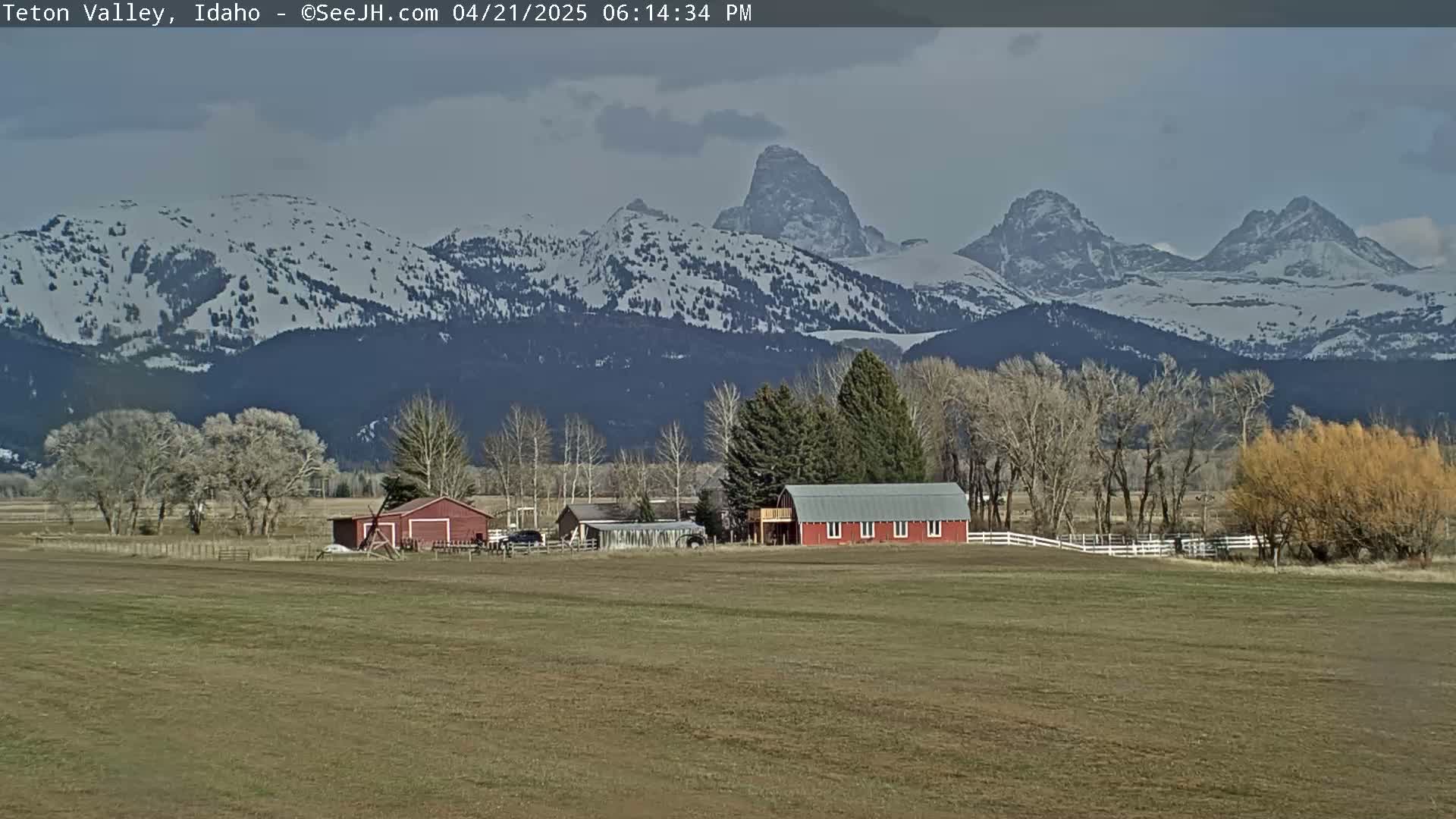 A red barn sits on a grassy field in front of snow-capped mountains under a partly cloudy sky.