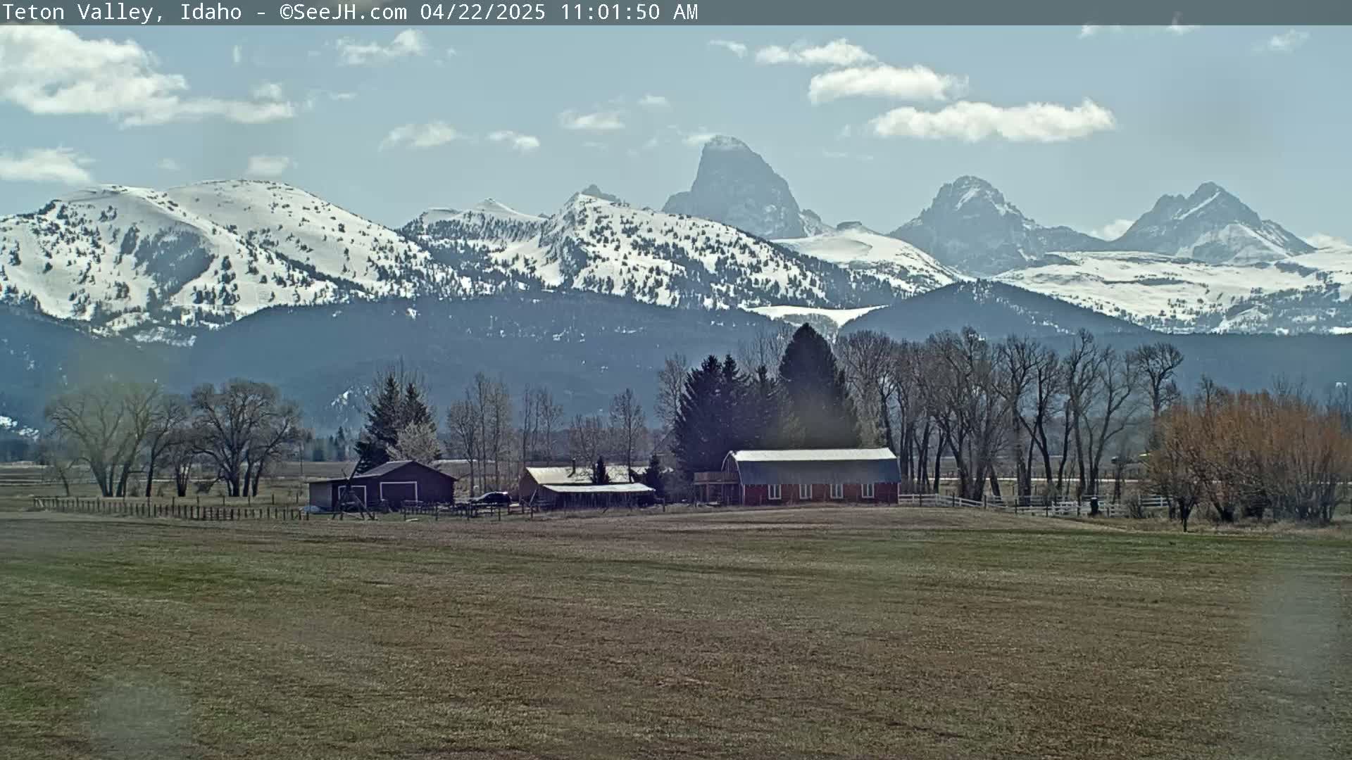 A farmhouse and outbuildings sit on a flat field in front of snow-capped mountains under a partly cloudy sky.