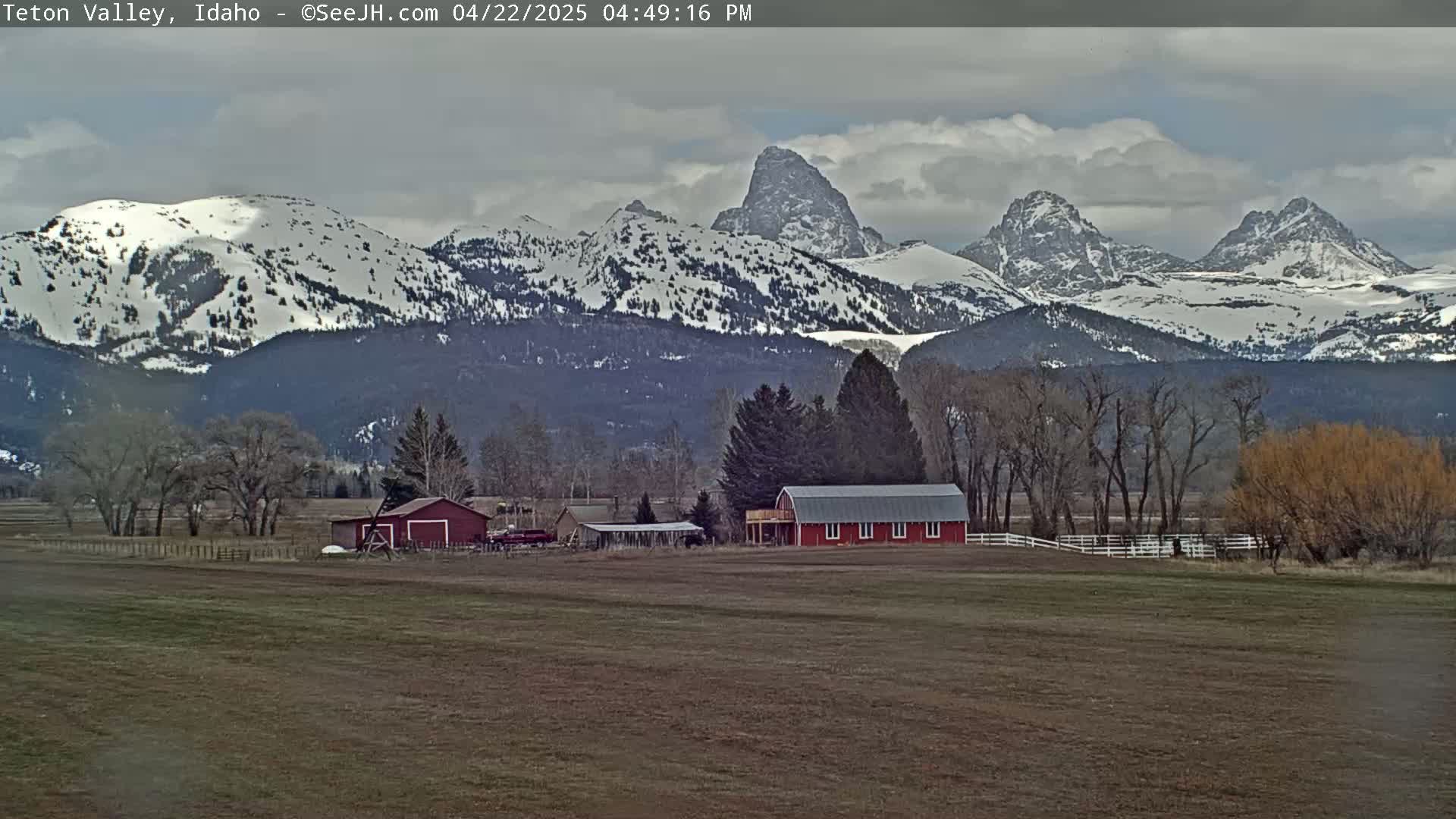 A partly cloudy day shows snow-capped mountains in the background, with a red barn and other farm buildings in a field in the foreground.
