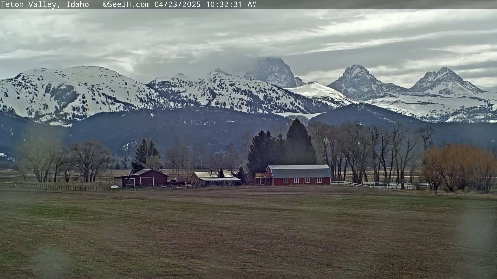 A red barn sits on a flat field in front of snow-capped mountains under an overcast sky.