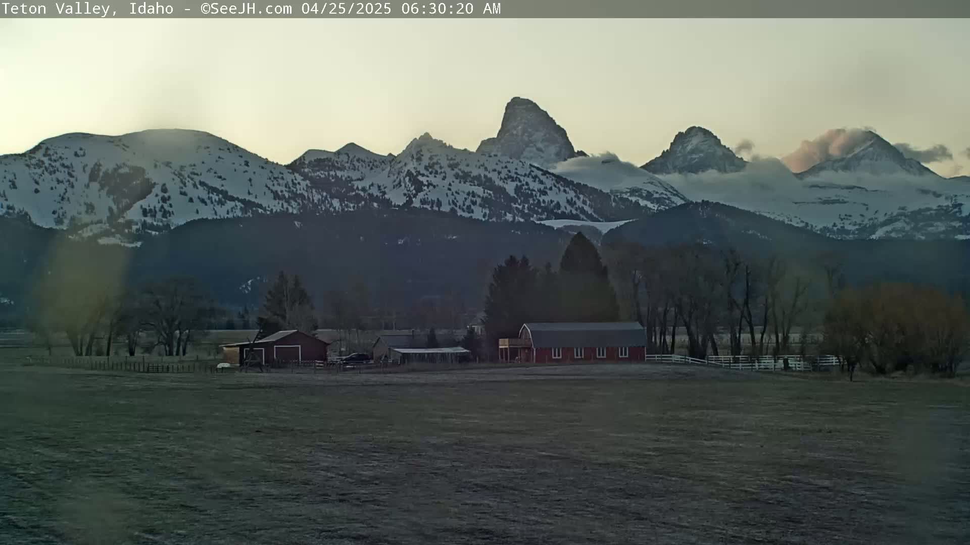 A red barn sits on a flat field in front of snow-capped mountains partially obscured by clouds at dawn.