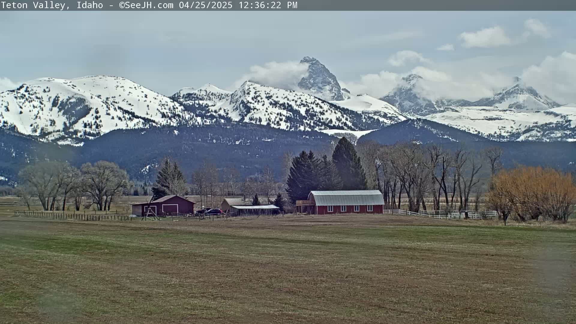 A partly cloudy day reveals a red barn and other farm buildings nestled in a valley before snow-capped mountains.