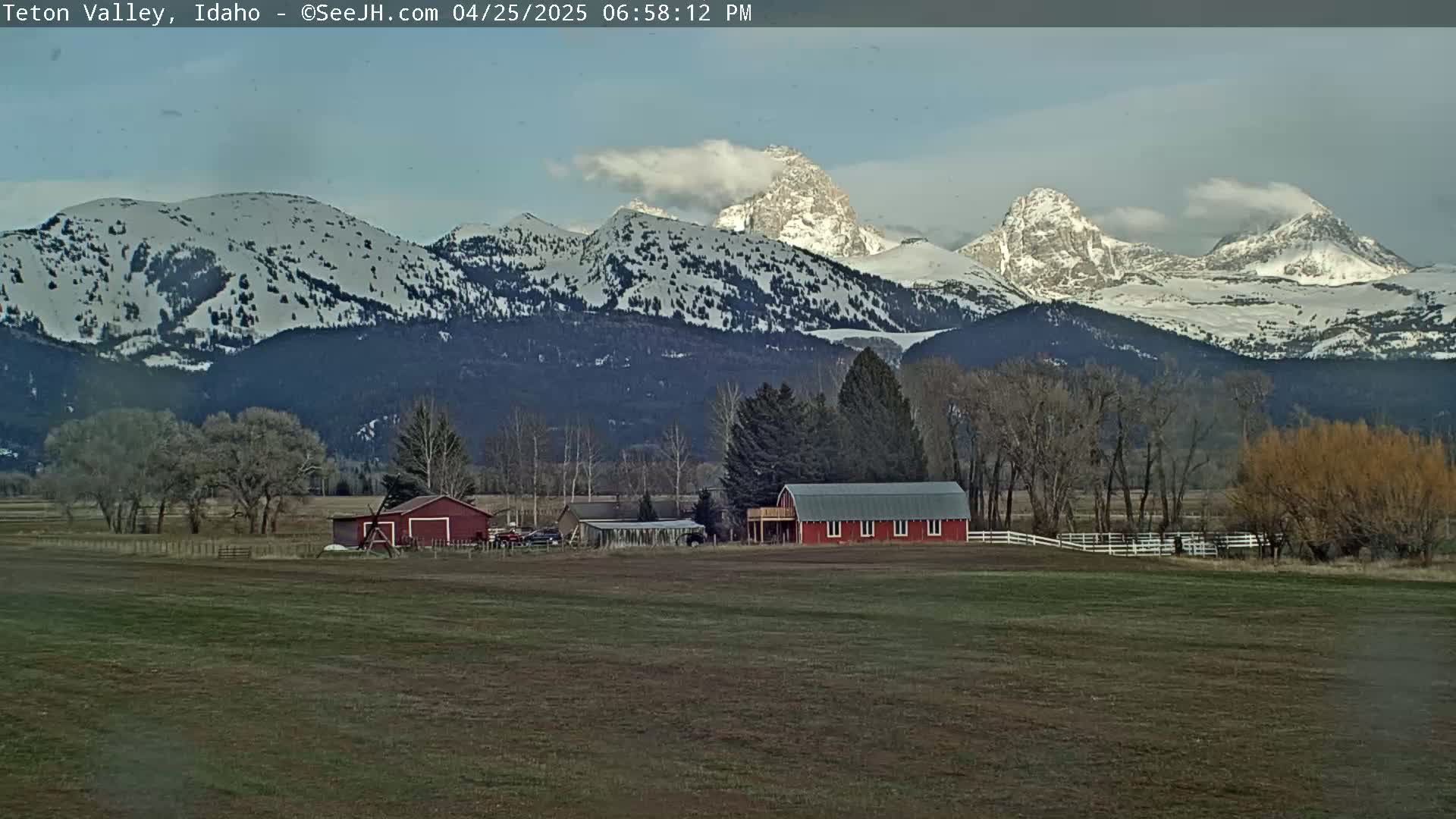 A red barn sits on a grassy field in front of snow-capped mountains under a partly cloudy sky.
