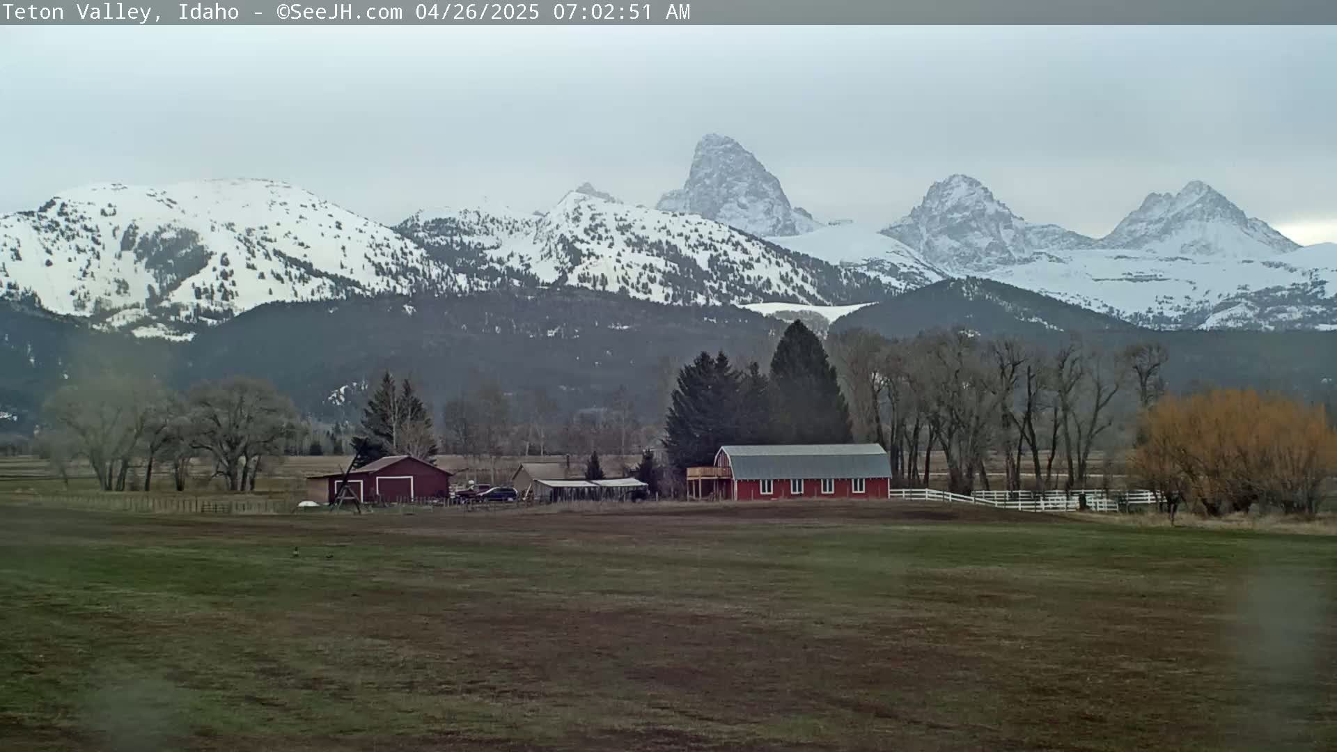 A red barn sits on a field in front of snow-capped mountains under an overcast sky.