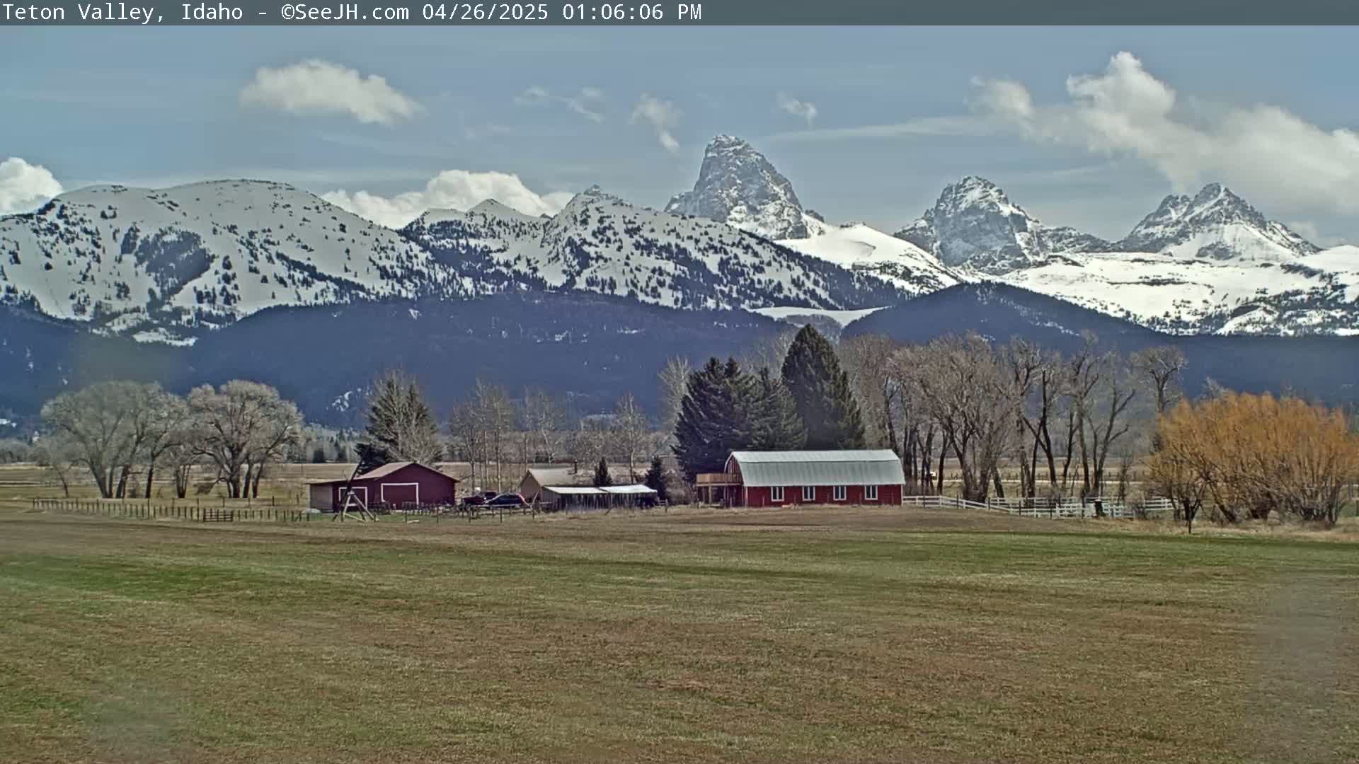 A red barn sits on a grassy field in front of snow-capped mountains under a partly cloudy sky.