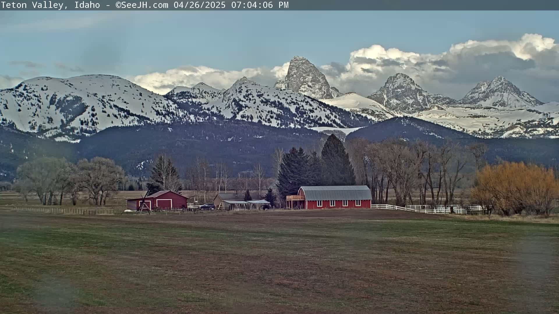 A red barn sits on a farm in a valley, with snow-capped mountains in the background under a partly cloudy sky.