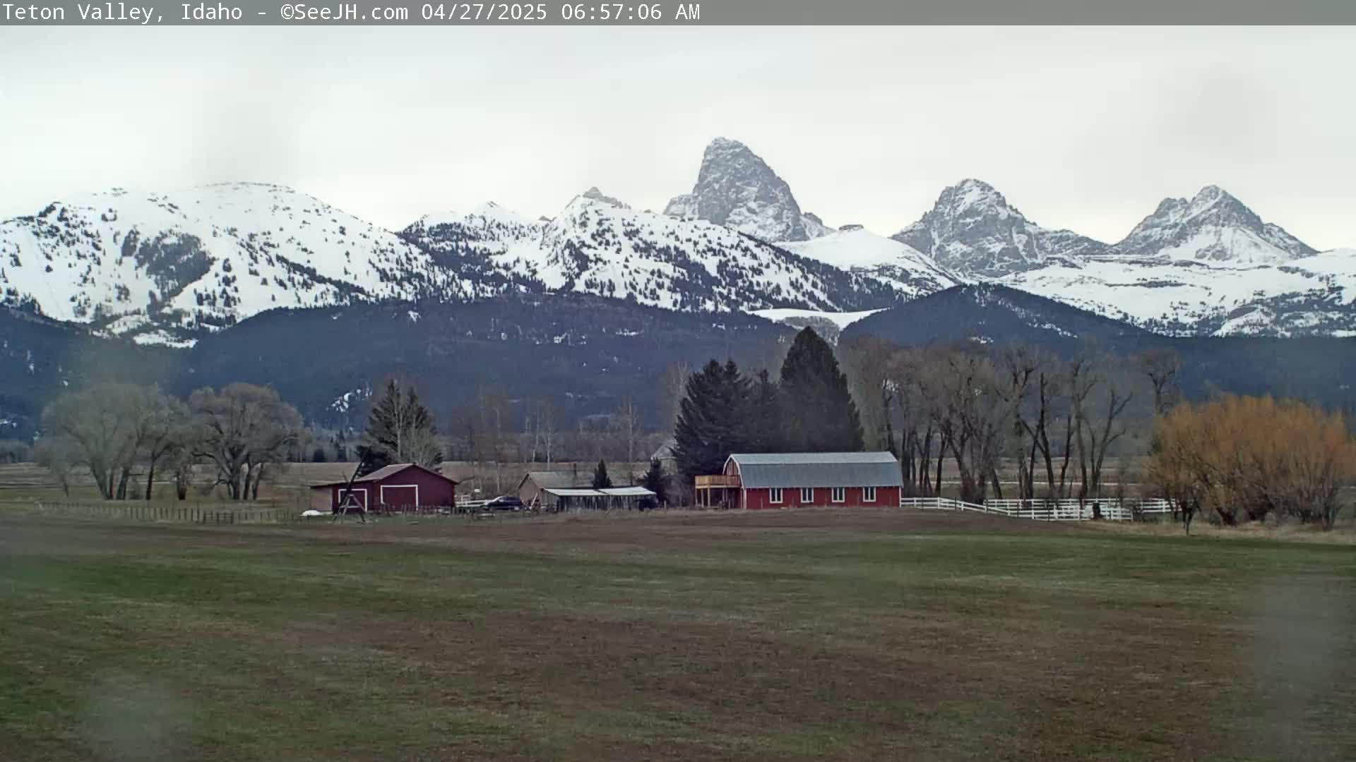 A red barn and other buildings sit on a flat, grassy field in front of snow-capped mountains under an overcast sky.